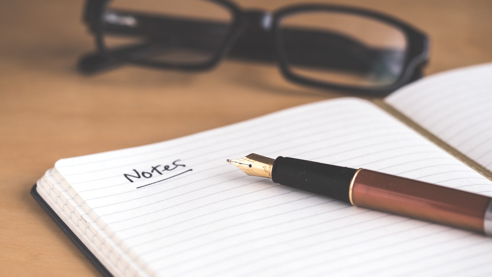 An open lined notebook with the word Notes handwritten and underlined, a fountain pen resting on the page and reading glasses behind it, August 2026