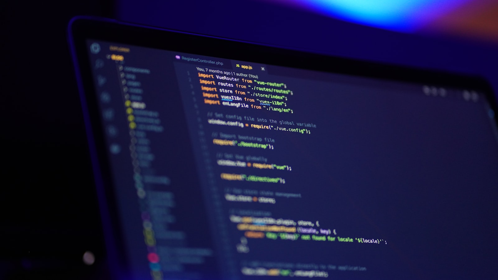 A laptop screen in a darkened room showing a code editor with a project file tree on the left and syntax-highlighted source on the right, lit in blue and purple, August 2026